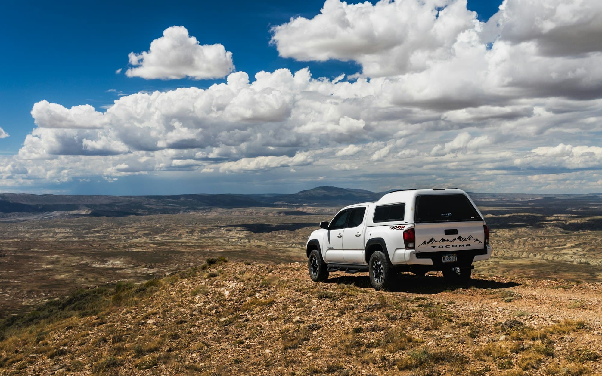 A white third-generation Toyota Tacoma with a bed cap parked above a wide desert valley